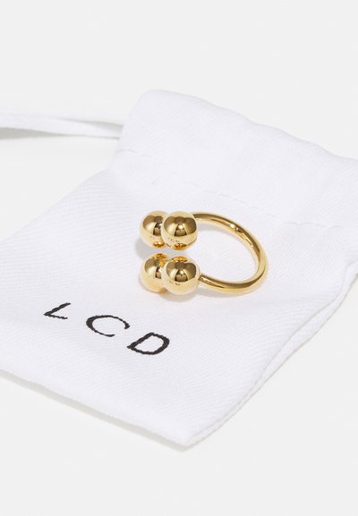 Gouden ring met drie sferische accenten, ingesteld op een dunne band. Gepresenteerd op een wit stoffen zakje met het label "LCD" in het zwart.