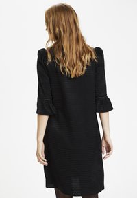 Svart klänning av texturerat tyg, 3/4-långa ärmar och volangärmar. Löst passande, knälång, minimal detaljering.