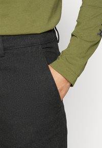 Camisa de manga longa verde-oliva combinada com calças de estilo denim escuras. As calças apresentam um bolso lateral e um tecido texturizado. Vista em detalhe do conjunto.