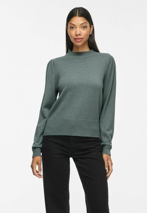 Femme aux longs cheveux foncés portant un pull vert à manches longues avec des épaules bouffantes et un pantalon noir, debout devant un fond blanc uni.
