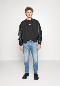 Zwarte bomberjack met satin mouwen, ritsluiting en geribde manchetten, gecombineerd met lichtblauwe jeans en zwarte laarzen.