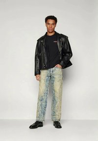 Juodas odinis motociklininko švarkas, juoda marškinėliai, šviesiai mėlyni blukinti džinsai su nusidėvėjusia tekstūra ir juodi batai. Modelis stovi prieš neutralią fono.