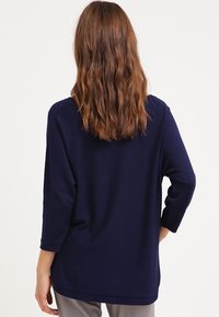 Námořnická modrá dlouhá rukávová blůza vyrobená z měkké tkaniny, s uvolněným střihem a kulatým výstřihem. Zpětný pohled ukazuje volný lem a texturovaný materiál.
