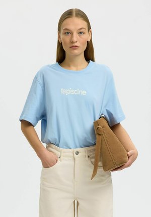 Mladá žena v svetlomodrom tričku s logom a krémových nohaviciach drží hnedú semišovú kabelku na jednofarebnom bielom pozadí.