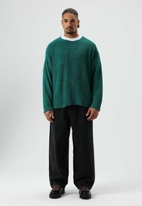 Turquoise, gebreide trui met lange mouwen en ronde hals, gecombineerd met losse zwarte broek en zwarte schoenen met dikke zolen en veters.