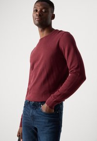 Red pullover sweater with long sleeves, ribbed neckline and cuffs, worn over blue denim jeans. Fabric appears smooth and fitted.