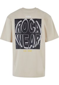 Bēšas krāsas īsām piedurknēm T-krekls ar lielu melnu grafiku, kurā redzams teksts "ROCA WEAR" un "EST. 1999" dzeltenā krāsā apakšā. Gluda auduma.