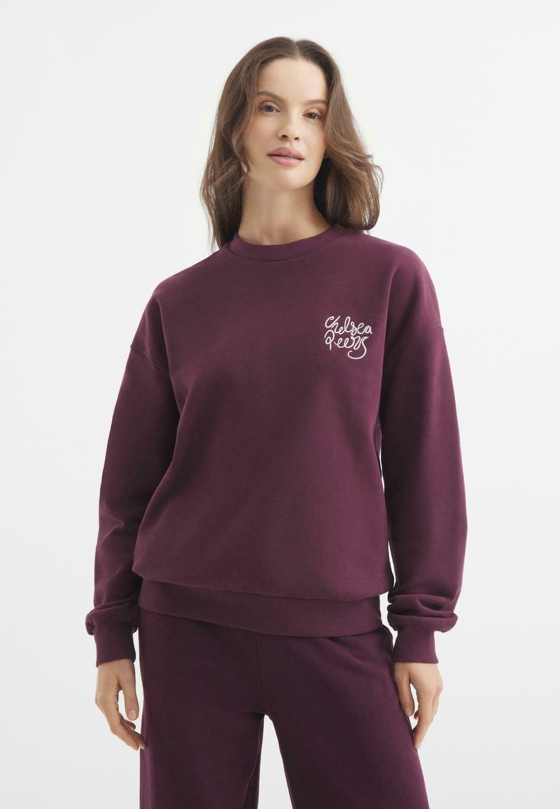 Sudadera burdeos de manga larga y cuello redondo. Presenta un logo blanco de "Chelsea Reess" en el pecho. Tejido suave y corte relajado.