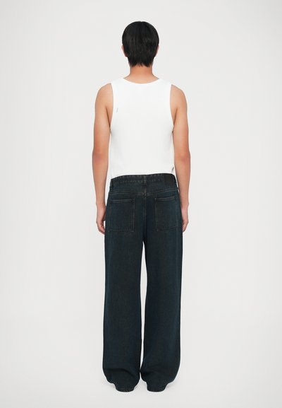 Άτομο με κοντά μαύρα μαλλιά, φορώντας ένα λευκό αμάνικο τοπ και φαρδιά σκούρα μπλε τζιν, που στέκεται και κοιτάζει μακριά σε έναν απλό φόντο.