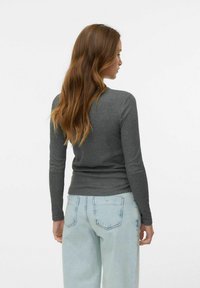 Femme avec de longs cheveux bruns ondulés portant un haut ajusté gris foncé à manches longues et un jean taille haute bleu clair, de dos.