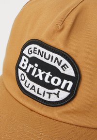 Brun hatt laget av teksturert stoff, med en sirkelformet svart og hvit lapp som står "Genuine Brixton Quality".