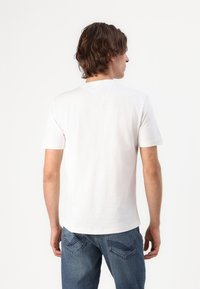 Camiseta blanca de manga corta con cuello redondeado, fabricada en algodón suave. Costura clara y una textura suave, combinada con jeans de mezclilla azules.
