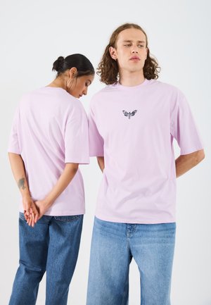 Lyserød bomulds t-shirt med korte ærmer og et centralt sort sommerfugl grafik. Modellen har afslappede blå denim jeans på.
