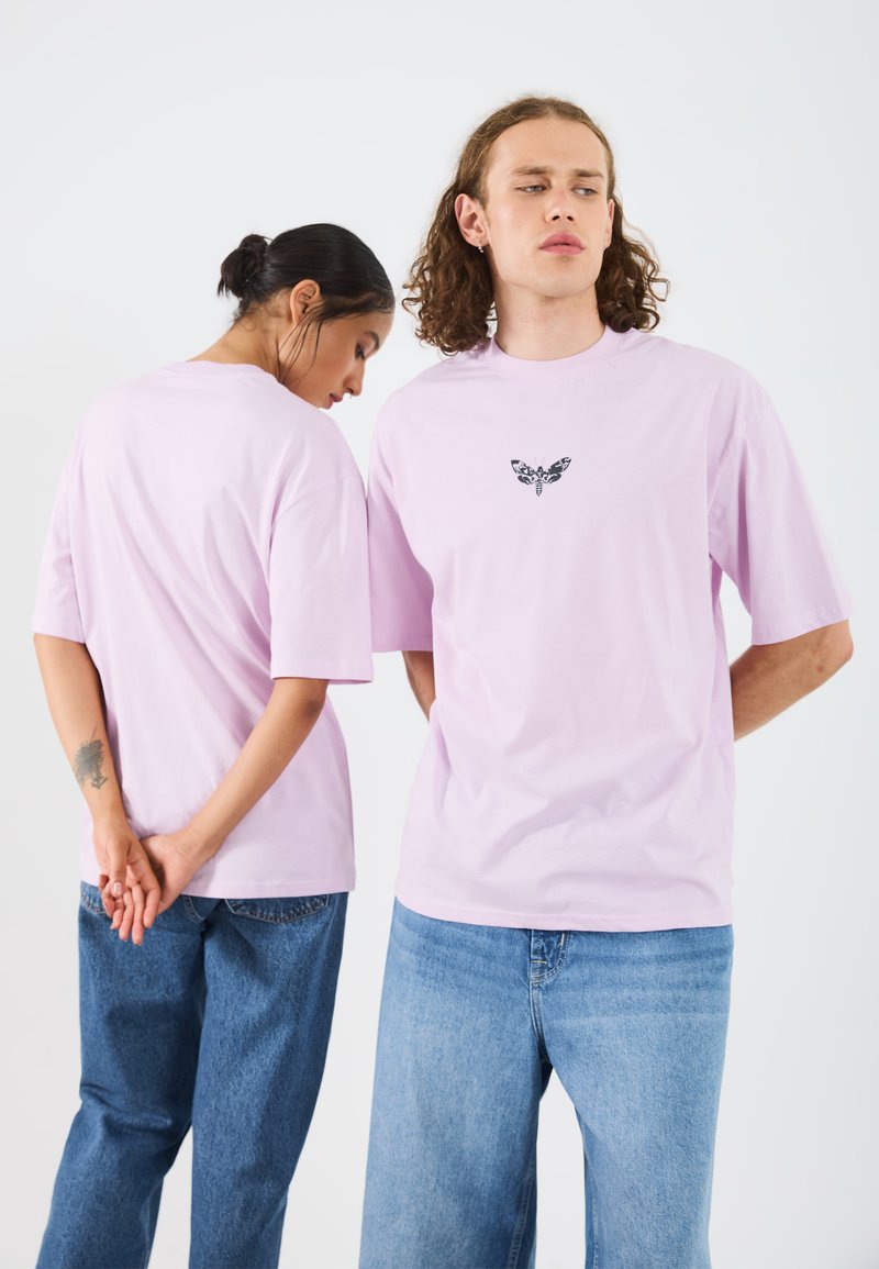 Light pink cotton t-shirt featuring short sleeves and a central black butterfly graphic. Model wears relaxed blue denim jeans.