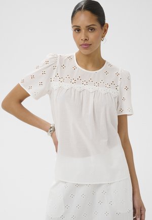 Žena v bielom krátkom tričku s krátkym rukávom a sukni s kvetinovými dierkovanými vzormi, stojaca s jednou rukou na boku pred jednoduchým pozadím.