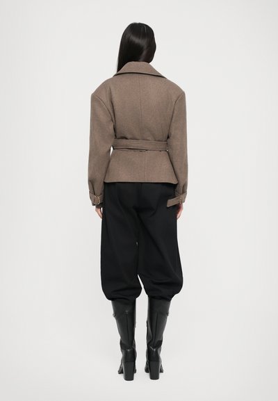 Brown fitted jacket with a belt, wide lapels, and structured shoulders, paired with loose black pants and knee-high black boots.