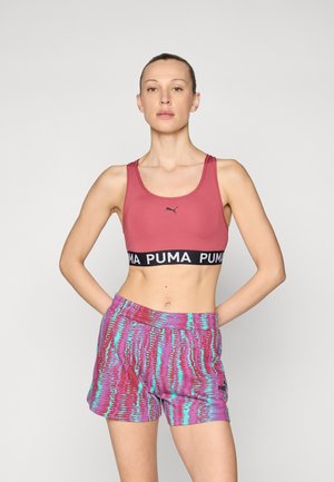 Biustonosz sportowy PUMA w kolorze różowym z czarną taśmą z logo, zestawiony z szortami w wielokolorowy wzór falisty w odcieniach purpury i turkusu.