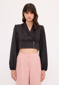 Schwarze Satin-Cropped-Bluse mit langen Ärmeln, V-Ausschnitt und drei goldfarbenen Knöpfen. Kombiniert mit hellrosa hochgeschnittenen Hosen.