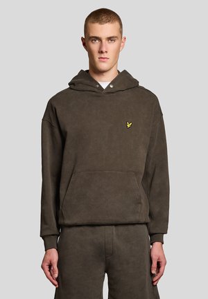 Jonge man met kort haar die een donkerbruin capuchontrui en bijpassende broek draagt, staand tegen een effen lichte achtergrond.