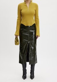 Kobieta w musztardowym, prążkowanym swetrze, błyszczącej czarnej skórzanej spódnicy midi z zamkiem błyskawicznym z przodu, czarnych, szpiczystych botkach i trzymająca musztardową kopertówkę.