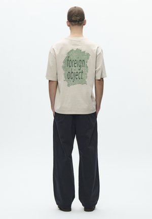 Muž v béžovom tričku so zeleným abstraktným dizajnom a textom "foreign object" na chrbte, v čiernych širokých nohaviciach a hnedej šiltovke, otočený chrbtom.