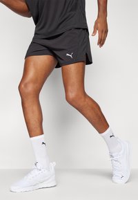 Svarta sportshorts med sidfickor och en liten vit Puma-logotyp, tillsammans med vita sneakers och vita ankelsockor med en logotyp.