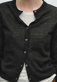Schwarze Strickjacke mit rundem Ausschnitt, ausgestattet mit sechs Knöpfen und einer leicht durchsichtigen Textur über einem weißen Hemd darunter.