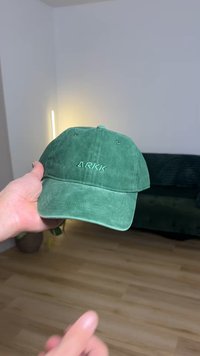 Casquette de baseball en daim vert avec le logo "ARKK" brodé. Visière arrondie, arrière ajustable, texture douce. En arrière-plan, un canapé et une source de lumière.