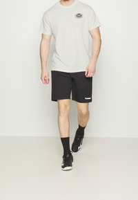 Světle šedé tričko s logem na hrudi, doplněné černými šortkami. Černé sportovní boty a černé kotníkové ponožky dotvářejí celkový vzhled.