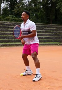 Tennis player wearing a white polo with gray and red stripes, pink shorts, and white shoes, holding a racket, with a yellow ball nearby.