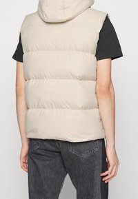 Beige pufferväst med huva, med kviltade sektioner och en slät yta, bärs över en svart t-shirt och matchas med mörka jeans.