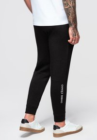 Czarne dresy o taperowanym kroju, wykonane z miękkiego materiału, z białym logo "OMBRE EXIMIO" na boku. Noszone z białymi i czarnymi trampkami.