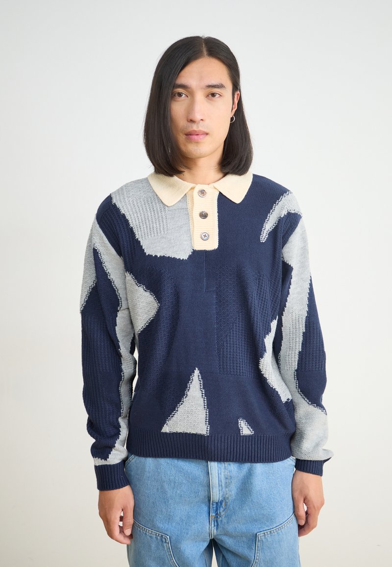 Πουλόβερ με μοτίβο σε ναυτικό μπλε, γκρι και κρεμ χρώμα; διαθέτει κολάρο, τρία κουμπιά και υφή πλεκτού υφάσματος.