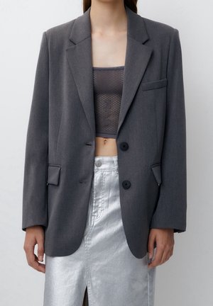 Vrouw draagt een oversized donkergrijze blazer over een grijs mesh crop top en een metallic zilveren hoge taille rok met een split aan de voorkant.