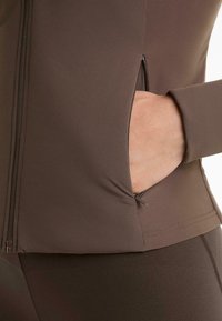 Rjav športni jopič z zadrgo na strani. Gladka tekstura, oprijet kroj in dolgi rokavi. Ujemajoče legging hlače.