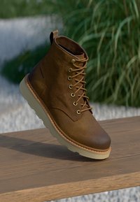 Bottine en cuir marron avec un bout rond, devant lacé et semelle en caoutchouc crème. Fini texturé avec un détail cousu autour de la semelle.
