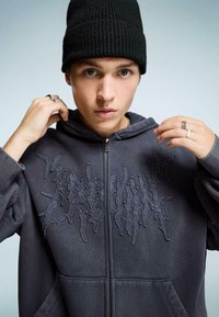 Temno siva zip-up kapuca z teksturiranim vezenim vzorcem spredaj, v kombinaciji z črnim rebrastim klobukom. Roke so vidne, nosijo prstane.
