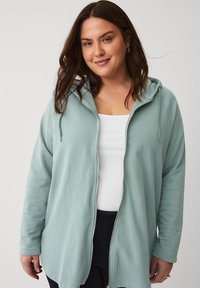 Sudadera de cremallera verde claro con capucha, mangas largas y un corte relajado, hecha de un tejido suave. Lleva puesta una camiseta sin mangas blanca debajo.