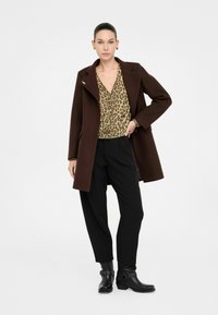 Cappotto marrone, su misura, con colletto alto, abbinato a una blusa con stampa leopardata e pantaloni neri. Stivaletti neri con fibbie completano l'outfit.
