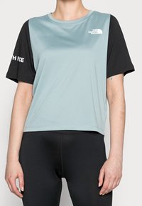 Osoba má na sobě sportovní tričko s krátkým rukávem, světle modré barvy s černými rukávy, doplněné černými legínami. Tričko je opatřeno logem "The North Face".