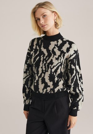 Donna bionda con un maglione fantasia astratta in nero e beige e pantaloni neri, in piedi contro uno sfondo chiaro e semplice.