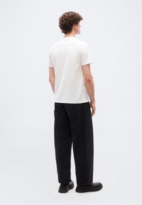 Άνδρας με σγουρά μαλλιά που φοράει απλό λευκό μπλουζάκι, άνετο μαύρο παντελόνι και μαύρα παπούτσια, στέκεται με την πλάτη γυρισμένη.