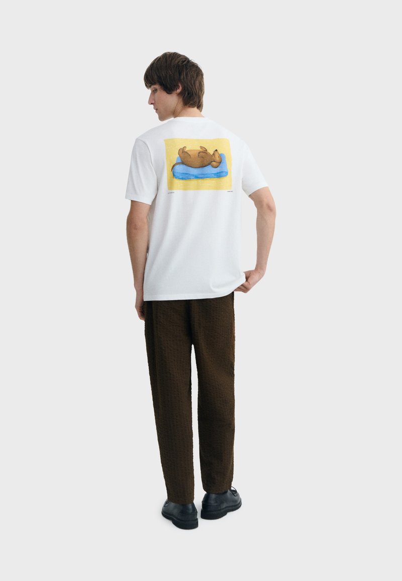 Jeune homme portant un T-shirt blanc avec un motif d'un chien marron couché sur un coussin bleu sur fond jaune, un pantalon marron et des chaussures noires.