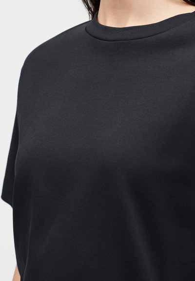 Γυναίκα που φοράει απλό μαύρο κοντομάνικο t-shirt με στρογγυλή λαιμόκοψη, εμφανίζοντας τον άνω κορμό και το λαιμό της μπροστά σε λευκό φόντο.