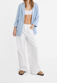 Hellblauer Blazer mit doppelreihigen Knöpfen, weißes kurzes Oberteil und weite, weiße Hose mit weitem Bein. Braune und schwarze Sandalen mit zwei Riemen.