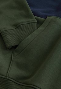 Sudadera de algodón verde con puños acanalados, que presenta una textura suave y un diseño en capas junto a una sección de azul marino oscuro.