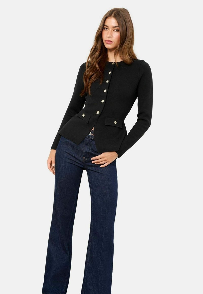 Cardigan negru cu un design ajustat, având nasturi aurii și două buzunare frontale. Asortat cu blugi evazați de culoare albastru închis.