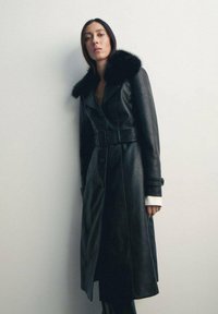 Trench coat in pelle nera con colletto di pelliccia, vita con cintura e chiusura a bottoni. Maniche lunghe con polsini, design strutturato.