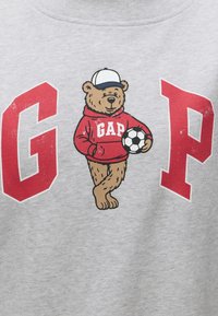 Ours portant un sweat à capuche rouge GAP et une casquette blanche, tenant un ballon de football, debout entre les grandes lettres rouges G et P sur un tissu gris.