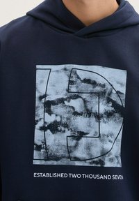 Granatowa bluza z kapturem z dużą, teksturowaną grafiką w odcieniach jasnoniebieskiego. Poniżej znajduje się napis "ZAŁOŻONA W DWA TYSCZE STOCZNE".